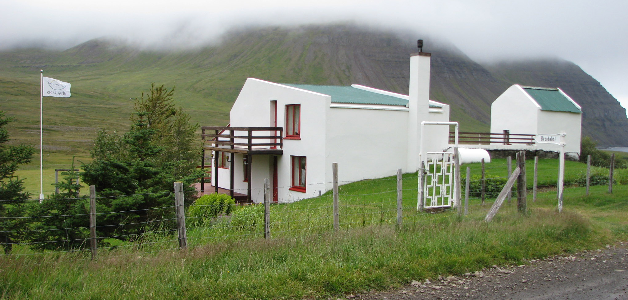 Breiðaból í Skálavík í júlí 2011. Öskubakur í baksýn, hulinn skýjum.  Áður var fleirbýlt á Breiðabóli en þetta hús er nú í eigu Karítasar Hafliðadóttur í Bolungarvík, ekkju Óskars Hálfdánarsonar, og afkomenda þeirra. Húsið stendur líklega þar sem Neðribær stóð, sem var eitt Breiðabólsbýlið, en þar bjó Jón faðir Jónu, Ingimundar, Sigurðar og þeirra systkina síðustu tvo áratugina. Breiðaból kemur víða við þessa sögu.
